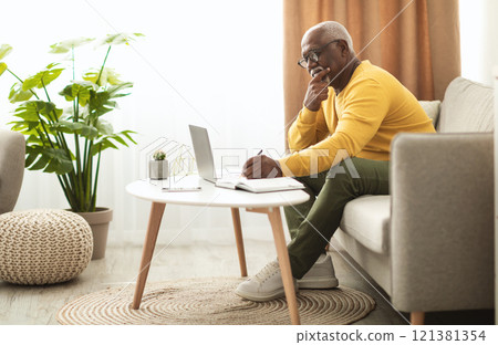 Mature African American Man Using Laptop Computer Taking Notes Working Online Sitting On Couch At Home. Senior Entrepreneur Doing Work In Living Room Indoor. Freelance Career. Side View 121381354