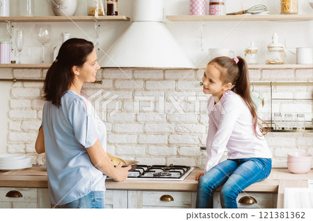 A woman and a young girl are cooking together in a modern kitchen. The woman is standing at the stove, stirring something in a pan, while the girl sits on the counter, watching her. 121381362