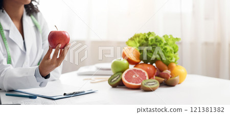 Healthy food concept. Cropped of nutritionist afro woman holding red apple in front of patient, consulting in clinic, panorama 121381382