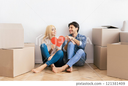 A couple sits on the floor of their new home, surrounded by boxes, holding a red heart. A couple sits on the floor of their new home, surrounded by boxes, holding a red heart. 121381409