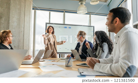 A businesswoman, standing in front of a whiteboard with a graph displayed, gestures towards the data as she presents information to a group of colleagues seated at a table in a modern office A businesswoman, standing in front of a whiteboard with a graph displayed, gestures towards the data as she presents information to a group of colleagues seated at a table in a modern office 121381500