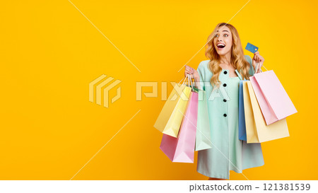 This image features a happy woman holding a stack of shopping bags and a credit card, against a bright yellow background. She is dressed in a light blue dress and has long blonde hair 121381539