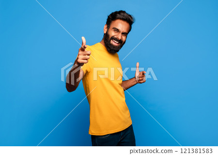 Indian man with a beard and a yellow t-shirt smiles and points at the camera with both hands. He stands against a blue background. 121381583