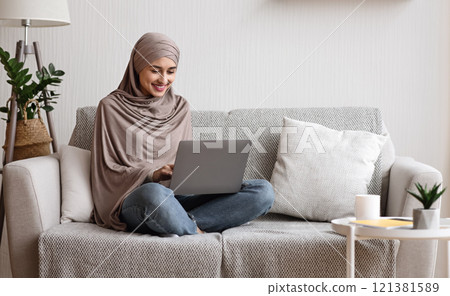 Modern arab girl in hijab using laptop at home, working remotely while sitting on couch in living room Modern arab girl in hijab using laptop at home, working remotely while sitting on couch in living room 121381589