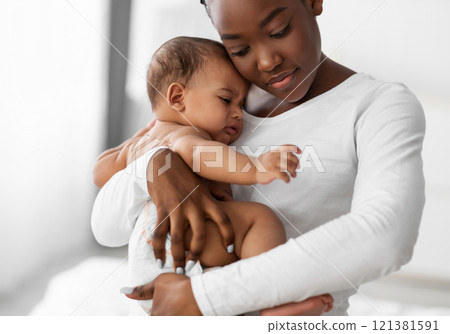 Maternity Concept. Portrait of young African American mother hugging and holding her upset crying baby on hands. Displeased hungry infant whining, feeling pain or colic. Copy space, blurred background 121381591