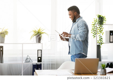 Successful african american entrepreneur holding digital tablet, browsing internet against window in modern office, side view Successful african american entrepreneur holding digital tablet, browsing internet against window in modern office, side view 121381621