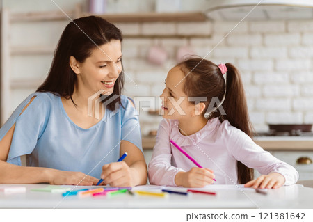 A mother and her young daughter sit at a table in their kitchen, coloring with colored pencils. They are looking at each other and smiling, enjoying a moment of bonding and creativity. 121381642