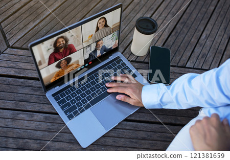 A person sits outdoors on a wooden patio, using a laptop to participate in a video conference. They are wearing a light blue shirt and white pants, top view 121381659