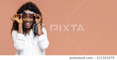 This image shows a Black woman with curly hair wearing sunglasses in a white shirt. She is smiling happily while posing against a light brown background. This image shows a Black woman with curly hair wearing sunglasses in a white shirt. She is smiling happily while posing against a light brown background. 121381679