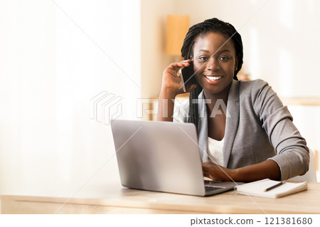 Smiling african american female manager talking on cellphone, consulting client, sitting at workplace in office Smiling african american female manager talking on cellphone, consulting client, sitting at workplace in office 121381680