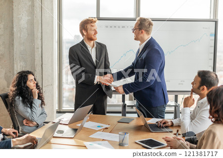 Two businessmen in suits are shaking hands in a modern office, while other colleagues look on. Large screen behind them displaying a graph, and laptops and other work materials are on the table. Two businessmen in suits are shaking hands in a modern office, while other colleagues look on. Large screen behind them displaying a graph, and laptops and other work materials are on the table. 121381717
