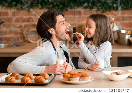 Caring little girl feeding her father with cookies while they having milk with snacks at kitchen, panorama with free space Caring little girl feeding her father with cookies while they having milk with snacks at kitchen, panorama with free space 121381747