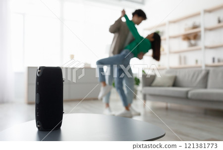 Millennial Asian couple dancing to popular music at home, selective focus on portable wireless speaker on table, copy space. Young family moving to their favorite song, using modern device Millennial Asian couple dancing to popular music at home, selective focus on portable wireless speaker on table, copy space. Young family moving to their favorite song, using modern device 121381773