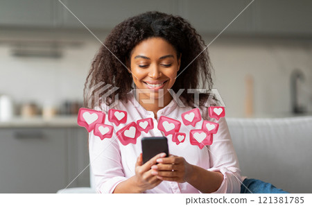Smiling black woman sitting on a couch and looking at her smartphone. Surrounding her are a number of pink heart-shaped icons, commonly used to represent social media likes 121381785