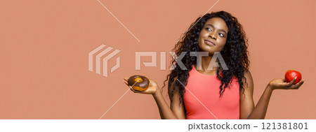 African American young woman with long curly hair, wearing a pink tank top, standing in front of a peach-colored background. She is holding a glazed donut in one hand and a red apple in the other African American young woman with long curly hair, wearing a pink tank top, standing in front of a peach-colored background. She is holding a glazed donut in one hand and a red apple in the other 121381801
