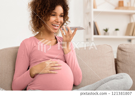 Happy pregnant woman sending voice message on phone, relaxing on sofa at home, free space 121381807