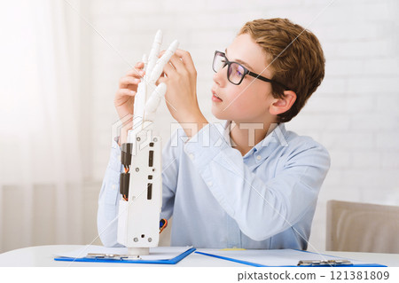 A young boy wearing glasses is sitting at a table in a classroom setting, intently examining a robotic hand model. He is holding the model and seems to be fascinated by its mechanics 121381809