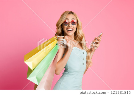 A happy blonde woman wearing sunglasses and a blue dress is holding a smartphone in one hand and multiple shopping bags in the other. She is standing in front of a pink background 121381815