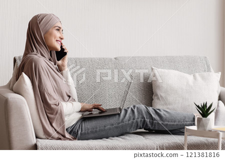 Relaxed muslim woman having phone call and using laptop while sitting on couch in a cozy apartment, side view Relaxed muslim woman having phone call and using laptop while sitting on couch in a cozy apartment, side view 121381816