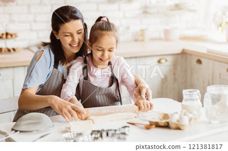 A mother and daughter work together in a kitchen, rolling out dough for cookies. The mother has her arm around the daughter and is guiding her hand as she uses a rolling pin 121381817