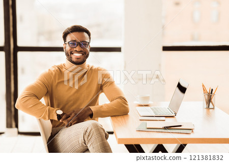 Enjoying Work. Smiling afro guy working on his laptop and looking at camera sitting at his working place, copyspace 121381832