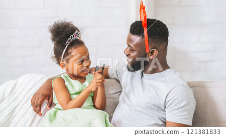 African American young girl wearing a crown and holding a makeup brush. She is sitting on the couch with her dad, who is wearing a red and orange headband. Both of them are smiling 121381833