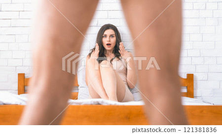 Woman seated on a bed, with her hands raised in a surprised and shocked expression. The legs of a man standing behind her are visible, suggesting a potential encounter or confrontation. Woman seated on a bed, with her hands raised in a surprised and shocked expression. The legs of a man standing behind her are visible, suggesting a potential encounter or confrontation. 121381836