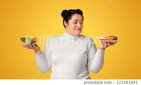 Uncertain plump girl can't decide between donuts and fresh vegetable salad, standing over ywllow background in studio with doubtful face expression 121381841
