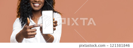 A young black woman with curly black hair is smiling and pointing at a blank smartphone screen. The image is great for showcasing a mobile app, website, or product, cropped 121381846