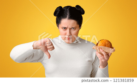 Stop Eating Fast Food. Annoyed overweight girl holding hamburger and showing thumb down, rejecting unhealthy meal, yellow background with free space 121381859