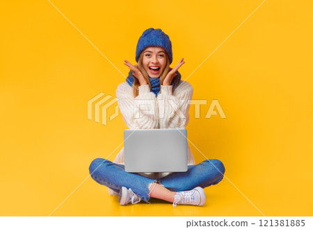 Big sales online. Beautiful young winter woman in knitted hat with laptop on her lap expressing happiness over pink background, sitting on floor 121381885