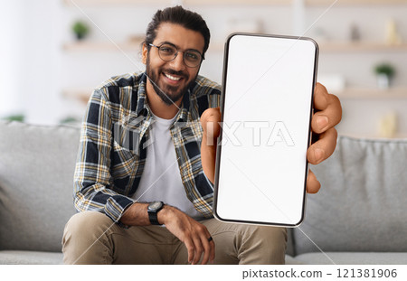 Cheerful Indian Guy Showing Smartphone With Big Blank White Screen At Camera, Happy Young Eastern Man Recommending New Mobile App Or Website While Sitting On Couch At Home, Creative Collage, Mockup 121381906