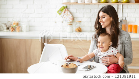 A woman is sitting at a kitchen table, holding a baby in her lap. She is working on a laptop computer, with a smartphone and a bowl of snacks in front of her, copy space A woman is sitting at a kitchen table, holding a baby in her lap. She is working on a laptop computer, with a smartphone and a bowl of snacks in front of her, copy space 121381911