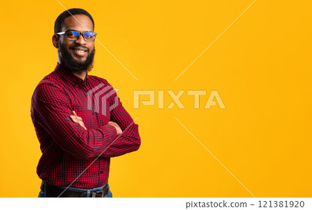 Confident Person. Portrait of smiling african american man wearing protective blue light glasses for computer, standing with folded arms isolated over yellow studio background wall with copy space 121381920