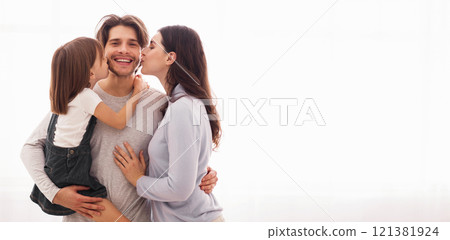 Sweetest kiss. Wife and daughter kissing happy man on both cheeks while standing together against window at home, copy space 121381924