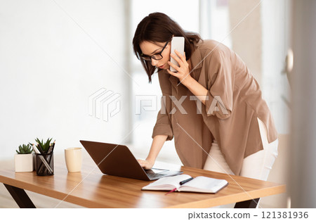 Business Person. Focused woman in glasses standing at desk using laptop computer and talking on mobile phone, copyspace 121381936
