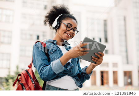 E-Learning. Happy Black Female Student Using Tablet Computer Wearing Headphones Standing Near Modern University Building Outside. Teenager Girl Browsing Internet Outdoors. Modern Online Education 121381940