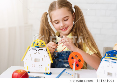 A young girl with pigtails is sitting at a table, intently building a robot toy. She is connecting wires and gears, showing focus and concentration as she works. 121381993