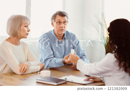 Medicine Concept. Female doctor giving bottle of medication to caucasian attentive senior couple, copyspace Medicine Concept. Female doctor giving bottle of medication to caucasian attentive senior couple, copyspace 121382009