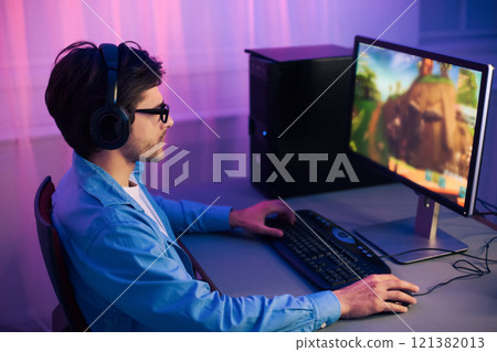 A young man, wearing headphones and glasses, is sitting in front of a computer monitor, playing a video game. The room is lit with purple lights, creating a vibrant and energetic atmosphere. 121382013