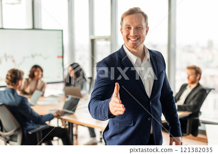 A smiling businessman in a blue suit extends his hand for a handshake while standing in an office, with other colleagues in the background. 121382035