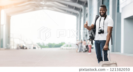 A happy black guy traveler, wearing a white shirt and carrying a backpack and suitcase, stands outside an airport terminal. He is smiling and giving a thumbs up as he walks towards the camera A happy black guy traveler, wearing a white shirt and carrying a backpack and suitcase, stands outside an airport terminal. He is smiling and giving a thumbs up as he walks towards the camera 121382050