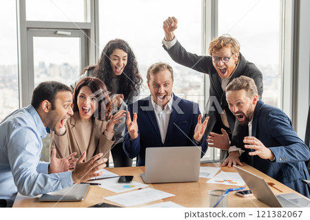 A diverse group of business professionals celebrates a successful project or achievement. They are gathered around a laptop, all wearing professional attire and beaming with joy 121382067
