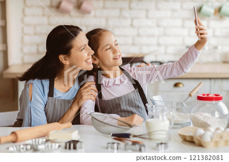 A mother and her young daughter are taking a selfie in the kitchen. They are both wearing aprons and smiling. There are baking supplies on the table in front of them 121382081
