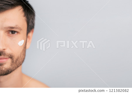 A man applies face cream to his cheek, looking directly at the camera. The image focuses on his face and features a simple, light-colored background. A man applies face cream to his cheek, looking directly at the camera. The image focuses on his face and features a simple, light-colored background. 121382082