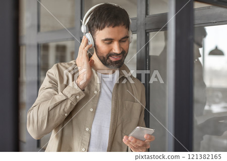 A man with a beard and short hair, wearing a beige button-down shirt and white undershirt, adjusts his headphones while holding a smartphone. He is standing in a modern office building 121382165