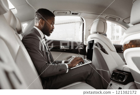 African American businessman sits in the backseat of a car, working on his laptop computer. He is wearing a suit and tie and appears to be focused on his work 121382172