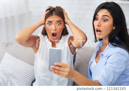 Omg. Shocked Girls Looking At Smartphone Screen Reading Message Sitting On Couch Indoor. Selective Focus 121382185