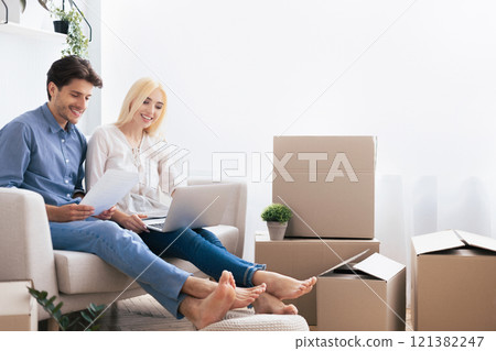 A young couple sits on a couch in their new home, surrounded by boxes. They are looking at a laptop and smiling. A young couple sits on a couch in their new home, surrounded by boxes. They are looking at a laptop and smiling. 121382247