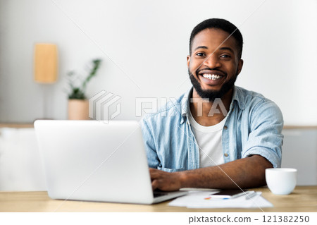 Portrait of handsome african worker using laptop, smiling and drinking coffee at workplace in modern office. Free space 121382250